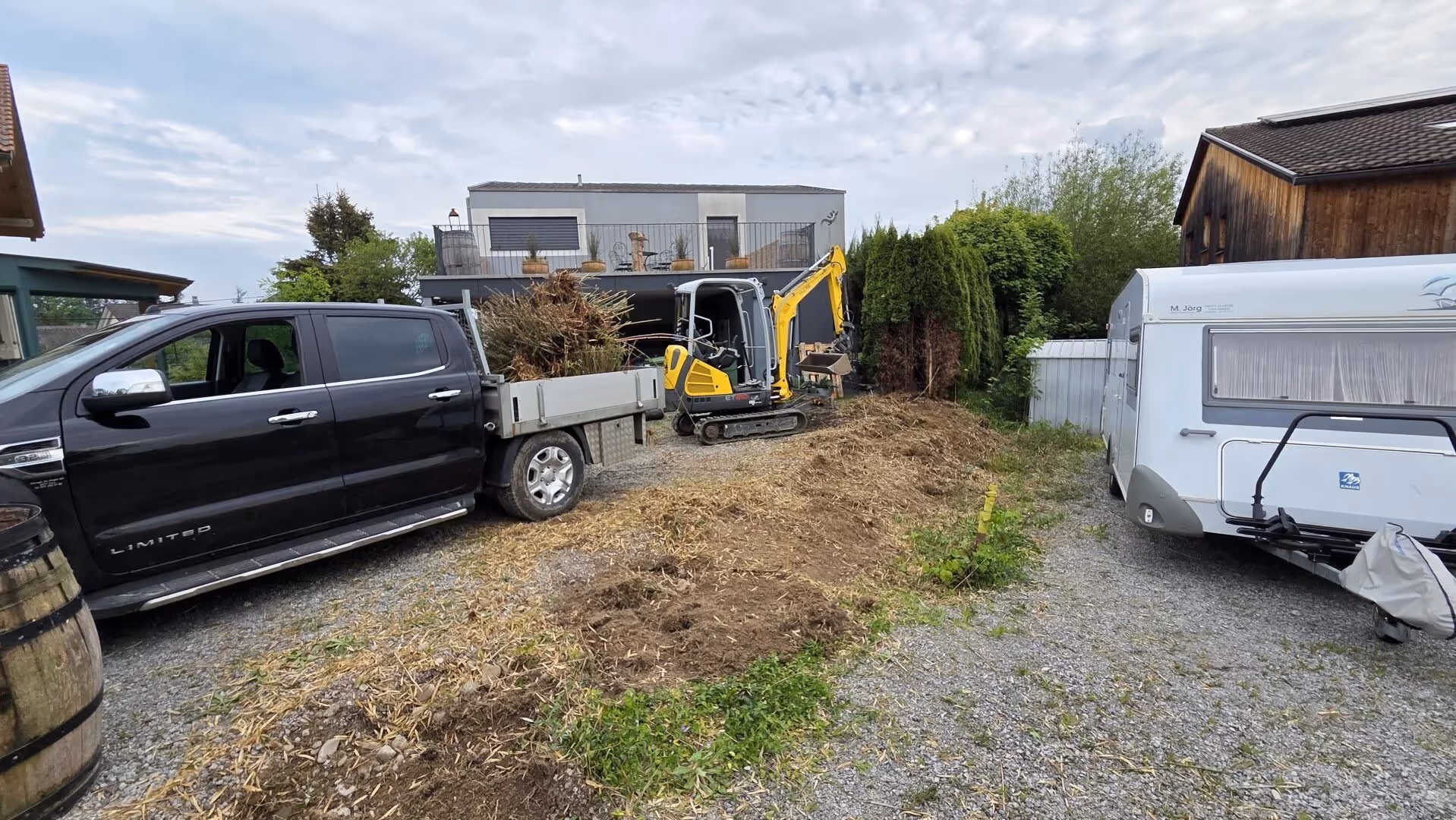 Schwarzer Pickup mit Ladung aus Ästen und Sträuchern neben gelbem Minibagger vor modernem Haus mit Terrasse und Wohnwagen auf Schotterweg.