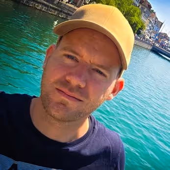 Mann mit gelber Kappe im Freien vor klarem blauem Wasser und Gebäuden am Ufer.